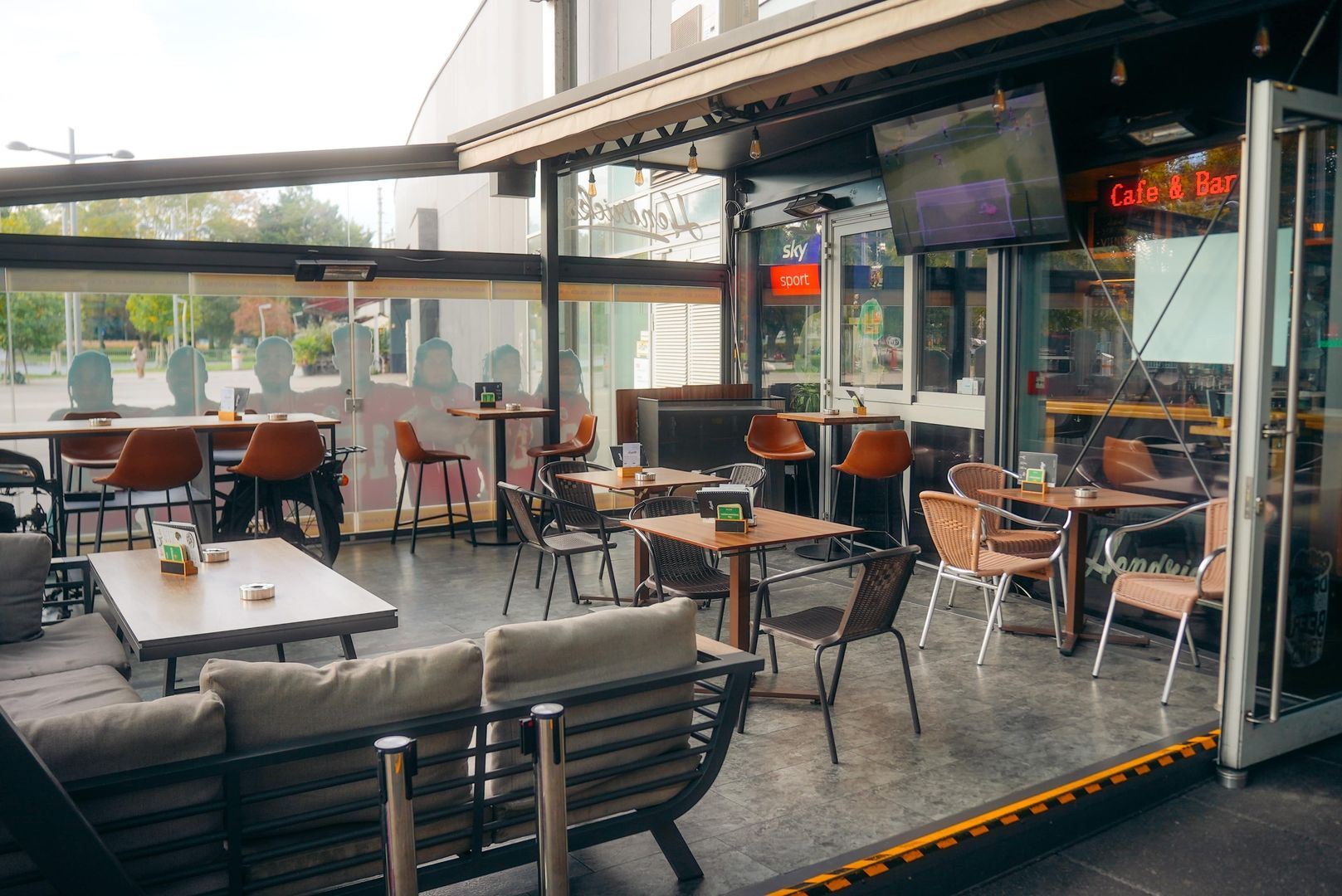 Conservatory view of Hendricks bar at Praterstern with comfortable seating, large windows, and multiple tables arranged for guests. A TV is visible in the corner showing a sports broadcast, emphasizing the bars sports-friendly atmosphere