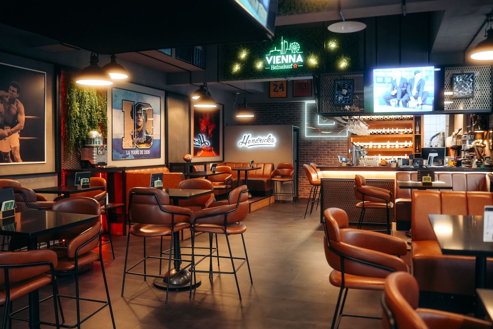 Interior view of the stylish Hendricks bar featuring cozy brown leather chairs, modern decorations, and atmospheric lighting. Sports and music-themed artwork can be seen in the background, along with a lit bar area.